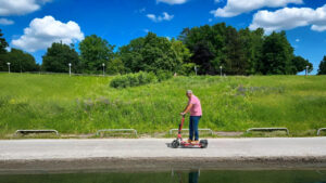 An old man rides a red e scooter for adults along a paved path beside a calm canal, with a grassy hill and trees in the background. He wears a pink shirt and jeans, enjoying a peaceful ride under a blue sky with white clouds.