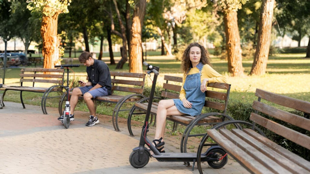 Young man and woman taking a break on park benches after riding e scooters for adults on a sunny afternoon in the park, surrounded by trees and quiet pathways.