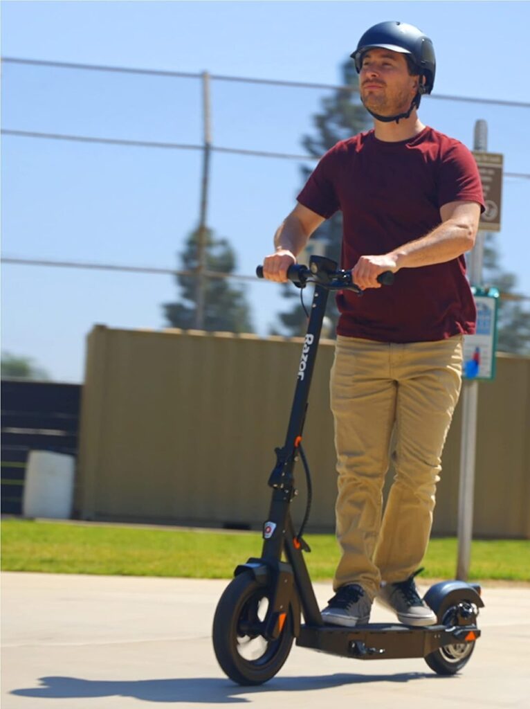 A man wearing a safety helmet rides a sleek black Razor electric scooter along a smooth pavement on a sunny day, with a fenced sports court and greenery in the background, confidently showcasing one of the best e scooters under 500$ for commuting, leisure rides, and daily urban travel.