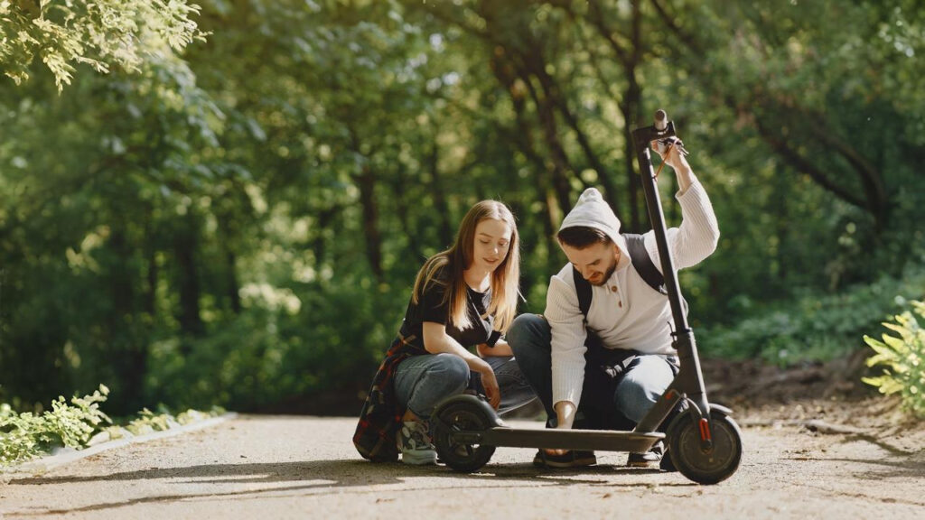 A young couple kneels on a sunlit forest path while examining the wheel of an electric scooter. The man holds the handlebar steady as the woman looks closely at the base of the scooter. Surrounded by lush greenery, they appear to be enjoying a peaceful outdoor moment while checking or fixing what could be the best e scooter under $500.