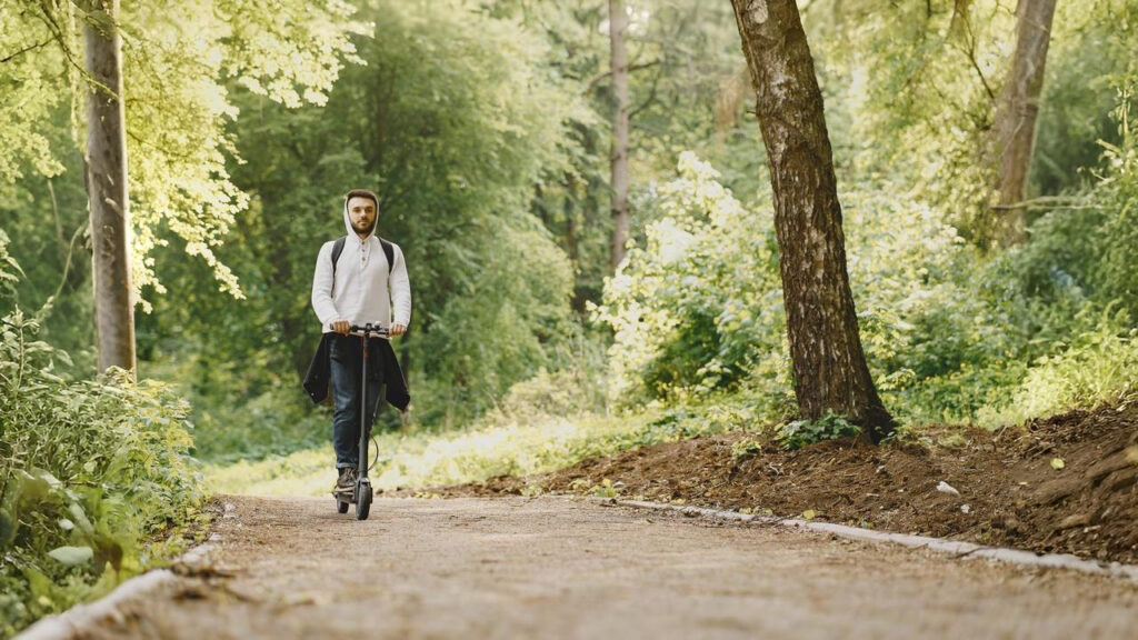 A man rides the best e-scooter for hills along a forest trail, wearing a white hoodie and backpack, highlighting powerful uphill performance, smooth handling, and reliability for outdoor commuting on uneven paths.