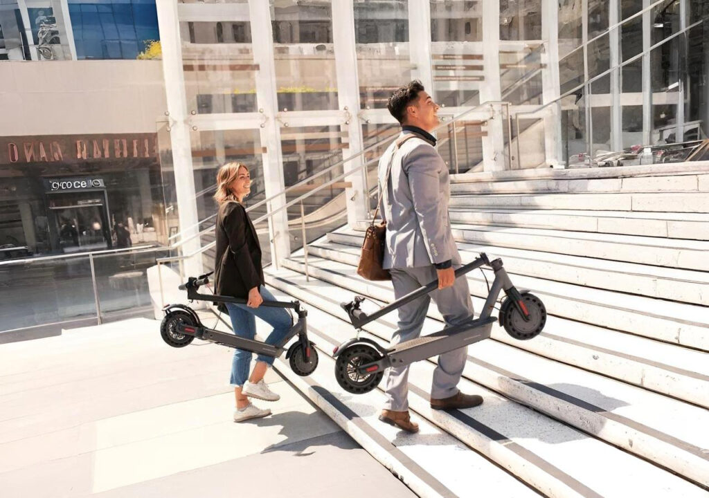 A man in a gray suit with a brown shoulder bag and a woman in a black blazer, jeans, and sneakers carry folding electric scooters up a wide set of modern outdoor stairs in a bright urban city environment.
