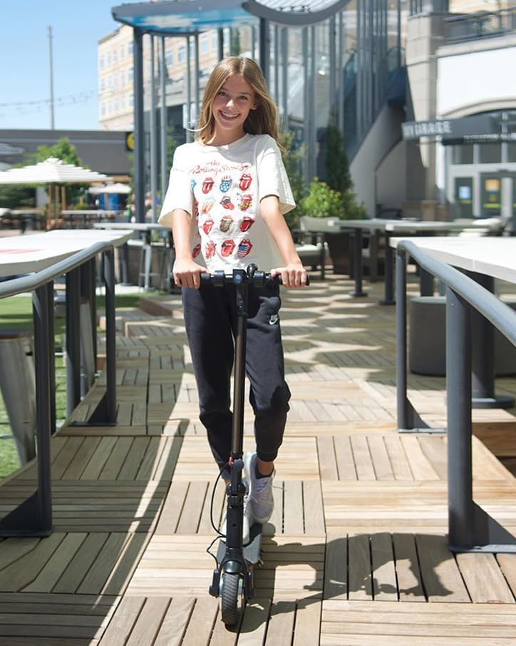 A young girl rides an electric scooter on a wooden walkway in a shopping area, enjoying a sunny day. 