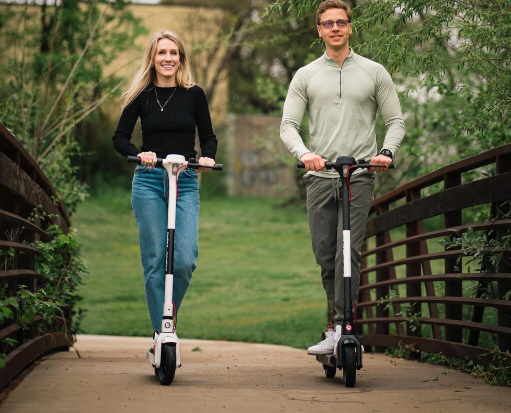 A man and a woman ride their electric scooters side by side on a bridge through a green park. These budget-friendly models are ideal for casual riders.