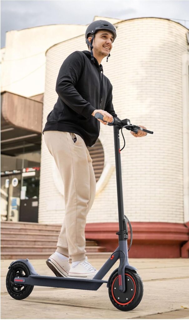 A man in a black hoodie and helmet rides an electric scooter with red accents near a modern building. He appears relaxed and happy, showcasing an affordable and reliable e scooter under $250 ideal for daily use.