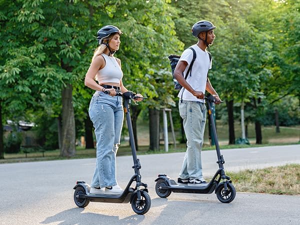 Man in white t-shirt and woman in white top riding NIU 1OOP electric scooters in a park, both wearing helmets and casual jeans.