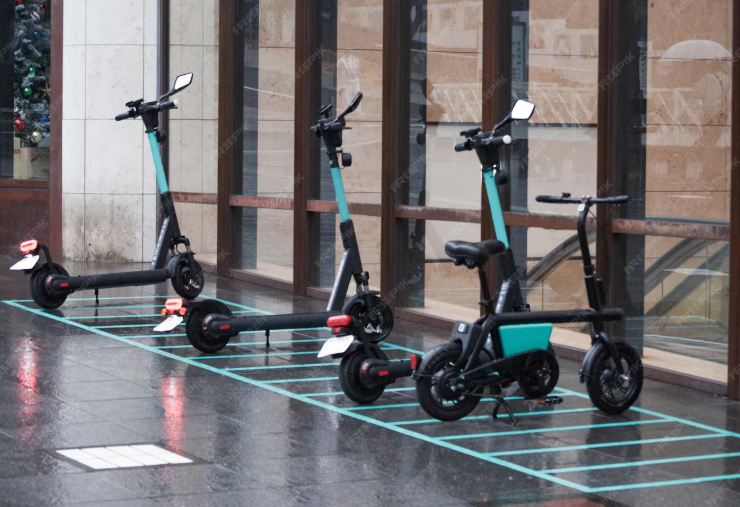 A row of electric scooters and a small e-bike parked neatly in a designated parking area with blue markings on a wet urban sidewalk in front of a modern building with large windows.