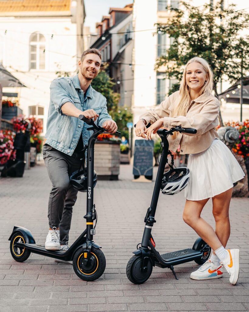 A smiling man and woman pose with their Atomi E30 electric scooters in a lively urban setting, highlighting eco-friendly commuting and modern city lifestyle.