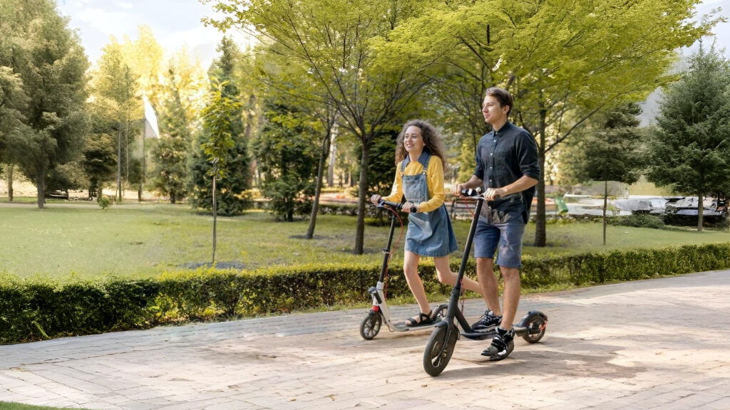 A young couple rides electric scooters through a park on a sunny day. The woman wears a yellow top with a denim dress, and the man wears a dark shirt with blue shorts. They both smile as they enjoy a leisurely ride on what could be the best electric scooter under $300, surrounded by trees and greenery.