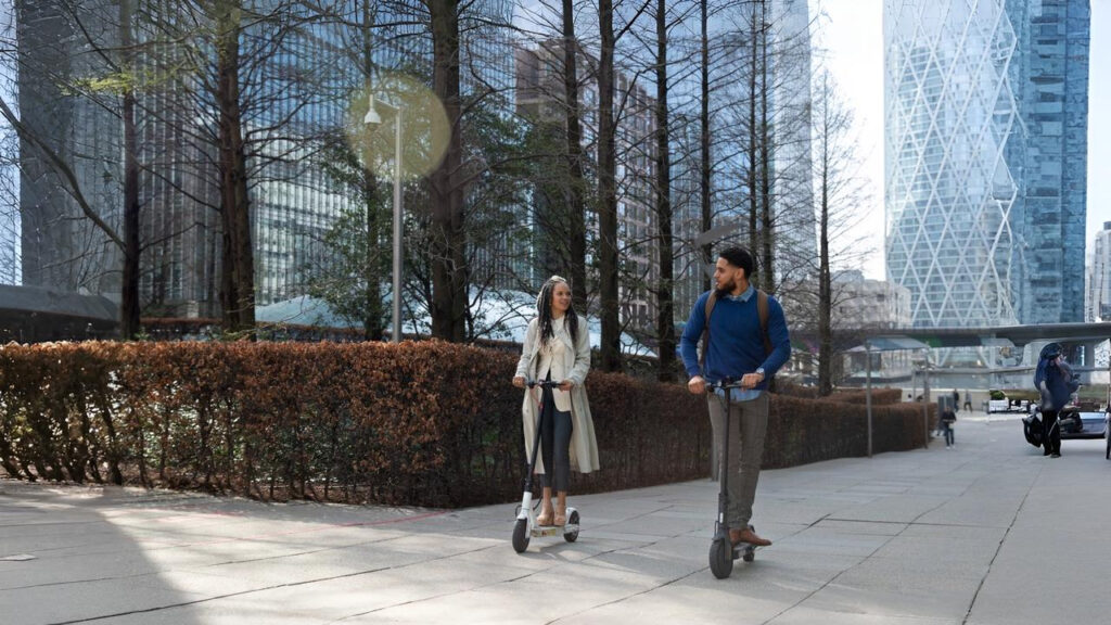 Two people ride electric scooters on a city pathway in NYC, surrounded by tall modern buildings, symbolizing urban mobility and raising the question: are electric scooters legal in NYC?
