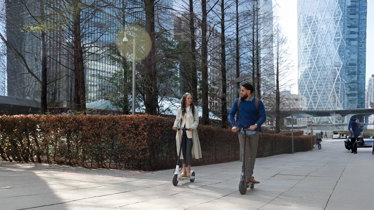 Two people ride electric scooters on a city pathway in NYC, surrounded by tall modern buildings, symbolizing urban mobility and raising the question: are electric scooters legal in NYC?