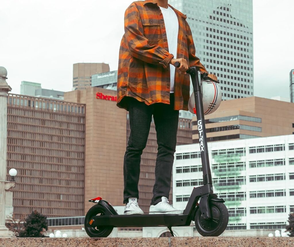 Alt Text: A person stands on a GOTRAX electric scooter in an urban setting, holding a helmet, with tall buildings in the background. This model is one of the best budget e scooters available for city commuting.