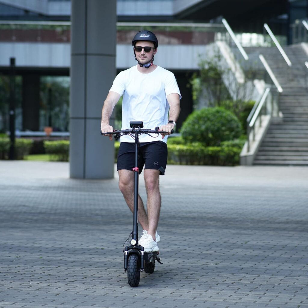 A man in a white t-shirt, black shorts, and sneakers rides the iScooter iX3 e scooter outdoors while wearing a black helmet and sunglasses