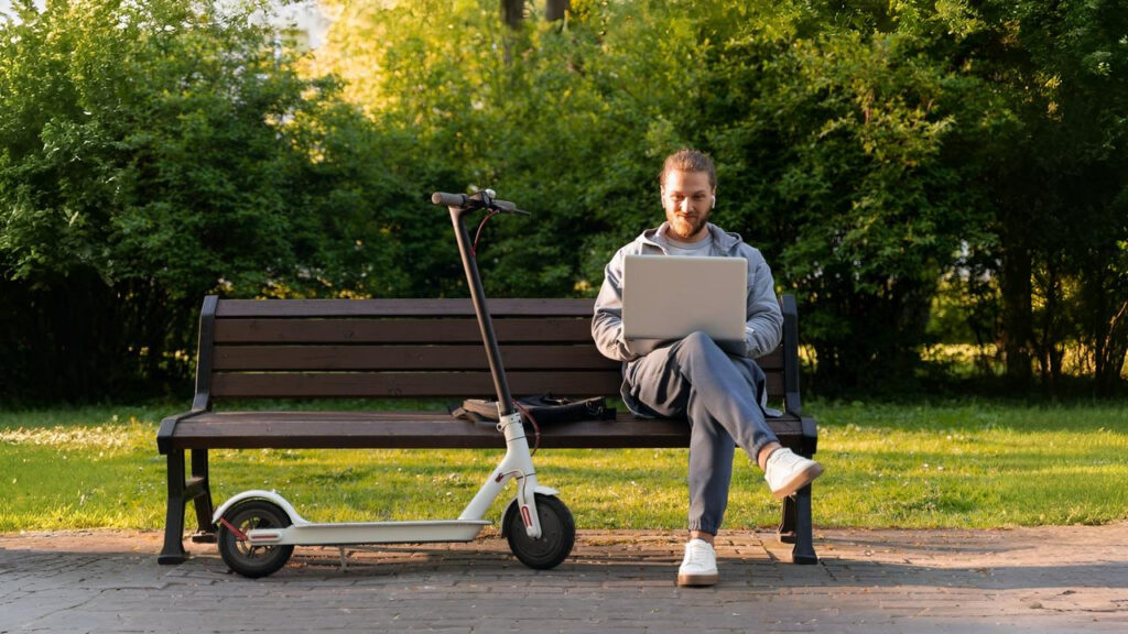 A man sitting on a park bench with a laptop beside one of the best budget e scooters parked next to him, enjoying remote work outdoors.