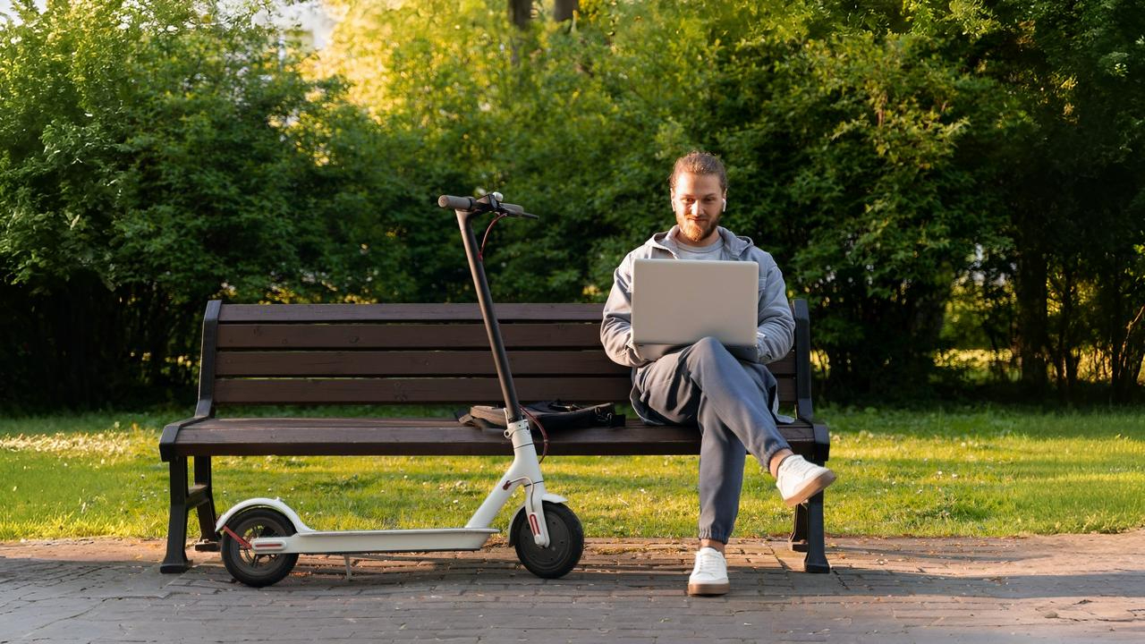 A man sitting on a park bench with a laptop beside one of the best budget e scooters parked next to him, enjoying remote work outdoors.