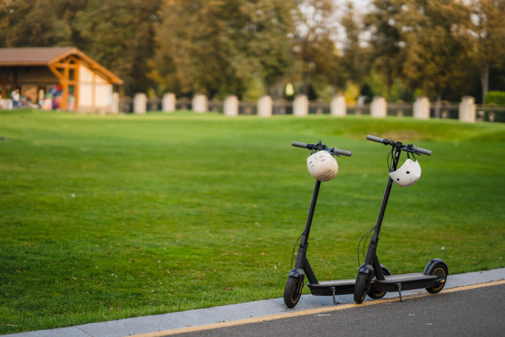 Two electric kick scooters with helmets hanging on the handlebars are parked on the edge of a paved path in Central Park, beside a large grassy area with trees and a wooden pavilion in the background.