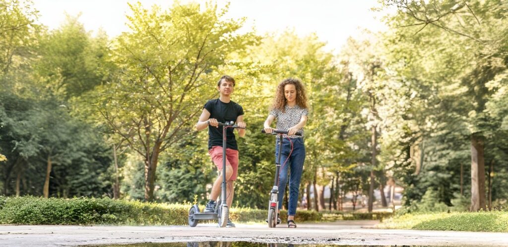 A young man in a black t-shirt and red shorts and a young woman in a patterned shirt and jeans are happily riding an e scooter under $250 on a sunny pathway in the park, surrounded by lush green trees and natural scenery, showing how affordable and eco-friendly scooters can be used for outdoor fun, daily commuting, and leisure rides.