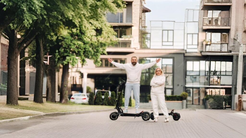 Two adults standing in the middle of a modern residential street with trees and contemporary buildings in the background, posing with two electric scooters. One man stands on a scooter with arms raised and smiling, while a woman stands beside another scooter with arms crossed, both wearing white outfits, showcasing the best brand e scooter lifestyle.