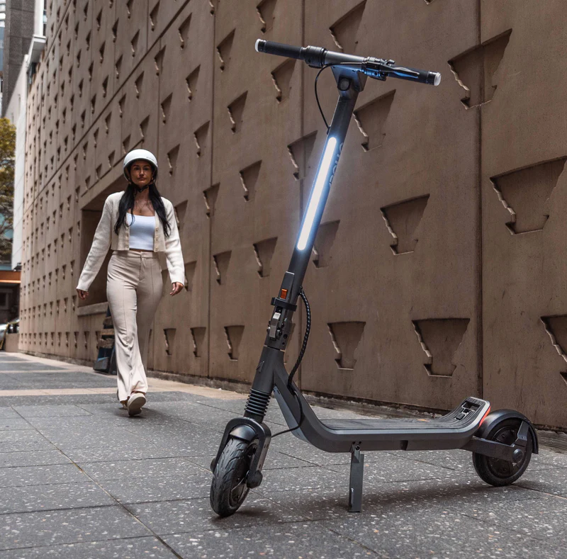 A woman wearing a helmet walks beside a parked e-scooter for heavy adults on a modern city sidewalk, with the scooter’s front light illuminated.