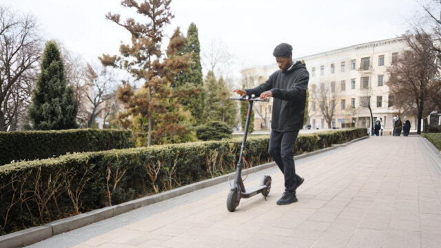 A man riding an e-scooter for heavy adults on a paved path in a park-like urban area, surrounded by trimmed hedges and tall trees.