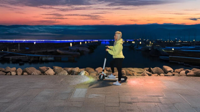 A woman riding an electric scooter along a waterfront promenade at sunset, with a marina and city lights in the background, showing the importance of LED lights for safety through visible front and rear lighting.