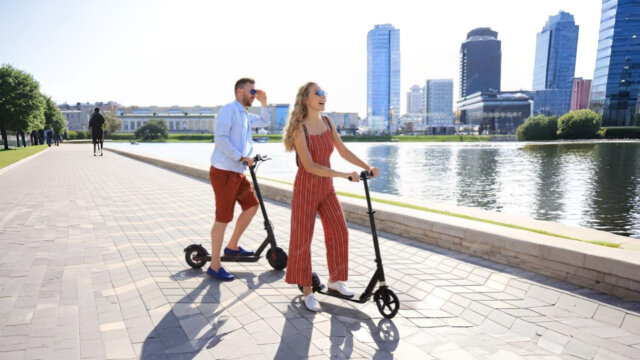 Two adults riding electric scooters along a waterfront promenade, representing eco-friendly transport options similar to the best budget e-bikes for city commuting.