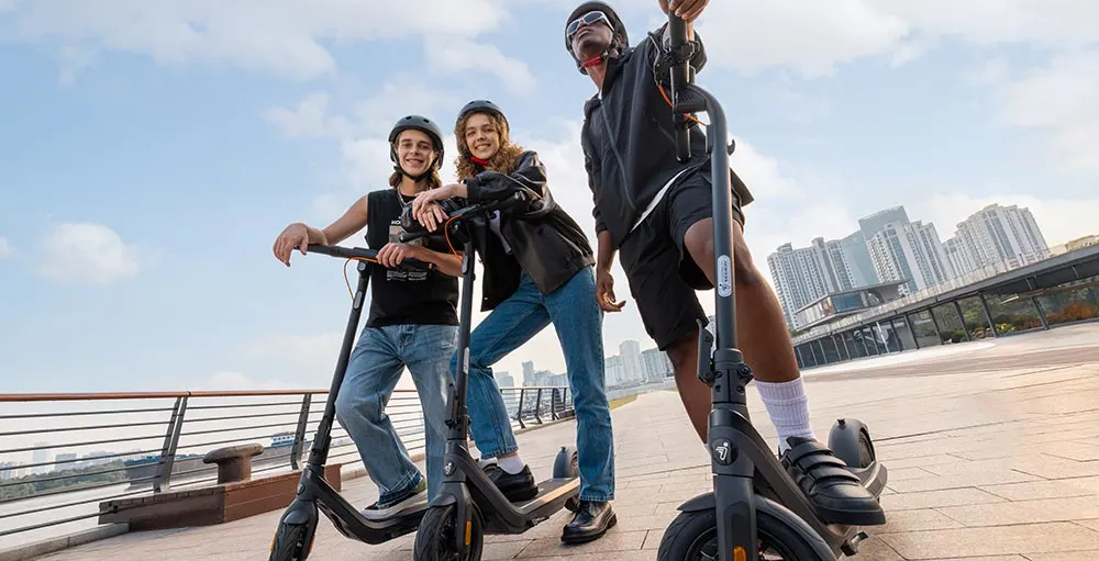 Three helmeted people pose with electric scooters on a waterfront promenade with city buildings in the background. The image suggests the best segway electric scooter for fun urban riding.