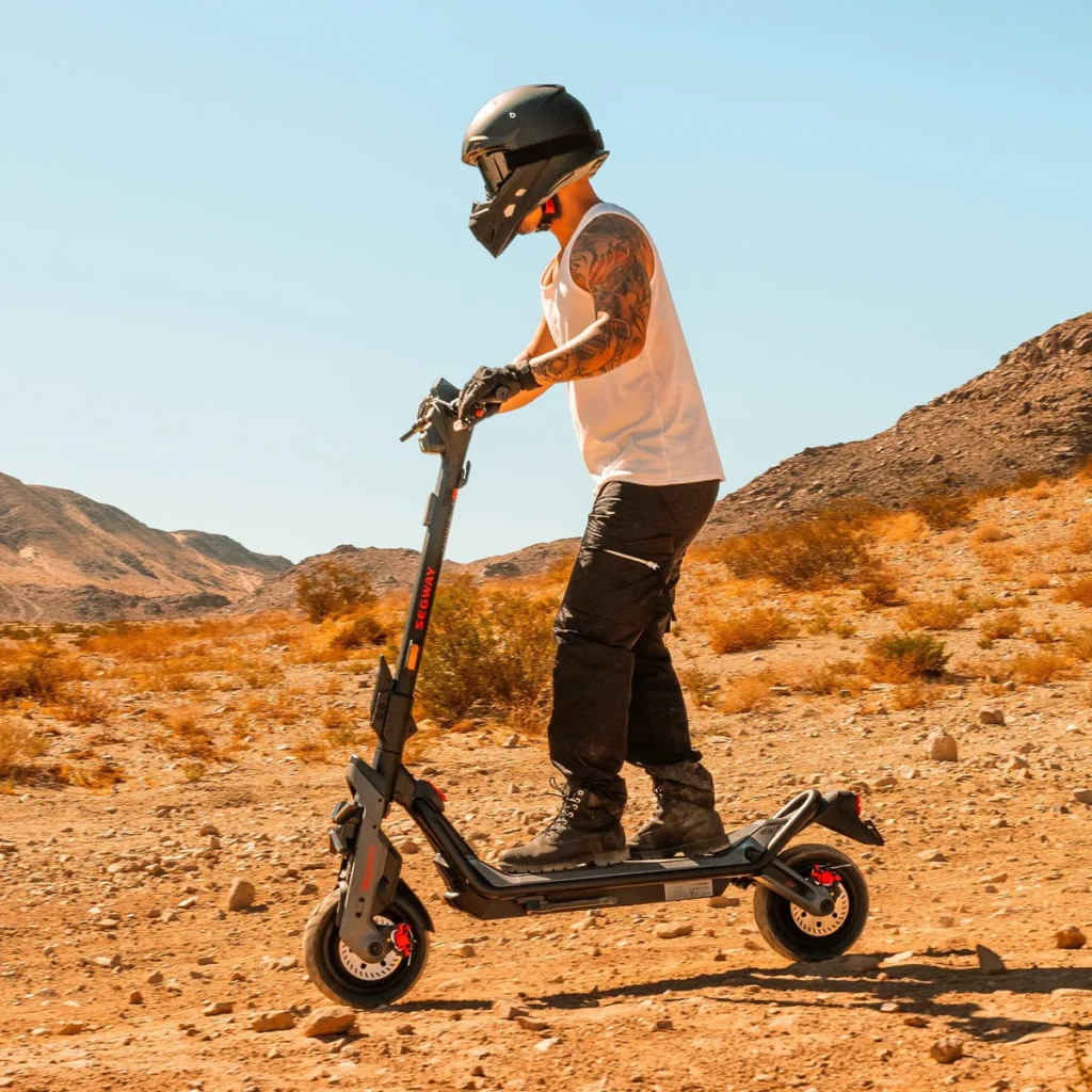Rider in a helmet and boots standing on a rugged Segway electric scooter in a desert landscape; an off-road best segway e scooter for adventurous riding.
