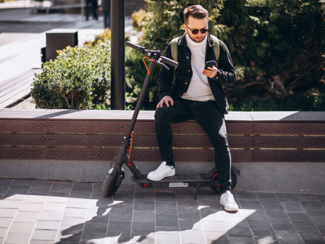 Man with sunglasses and backpack sitting on a parked scooter, looking at his phone outdoors in sunlight; a best lightweight e scooter is visible beside him.