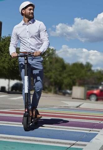 A man in a white shirt and helmet rides a scooter across a colorful painted crosswalk on a sunny day, demonstrating the best lightweight e scooter for smooth and fun city travel.