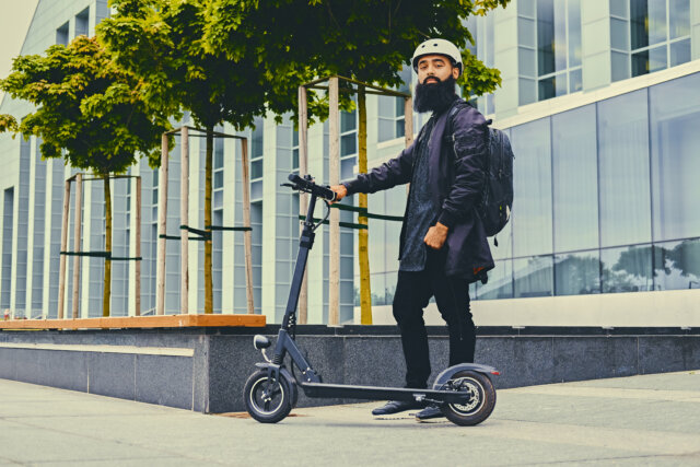 A stylish bearded man wearing a helmet and sunglasses poses beside an electric scooter on a city sidewalk, with modern glass buildings and trees in the background. Best Lightweight Electric Scooters